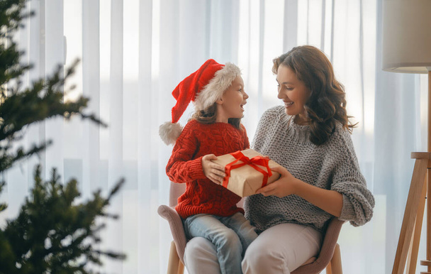 Merry Christmas, Happy Holidays. Cheerful mom and her cute daughter girl exchanging gifts. Parent and little child having fun near tree indoors.   - Photo, image