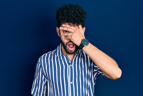 Young arab man with beard wearing casual striped shirt peeking in shock covering face and eyes with hand, looking through fingers afraid  - Fotó, kép