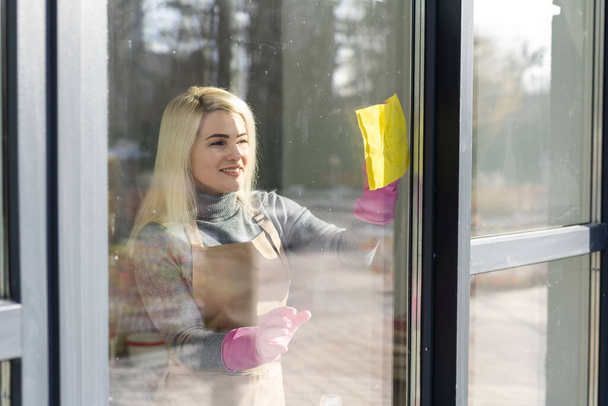 Νεαρή γυναίκα με ποδιά καθαρίζει παράθυρα. Υπηρεσία καθαριότητας. Καθαρισμός καμαριέρας στο σπίτι - Φωτογραφία, εικόνα