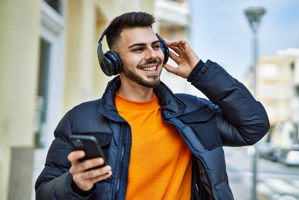 Handsome hispanic man with beard smiling happy and confident at the city wearing winter coat listening to music from smartphone - Foto, Imagem