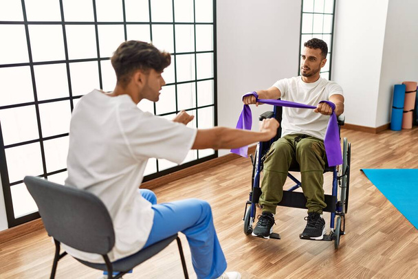 Two hispanic men physiotherapist and patient sitting on wheelchair having rehab session using elastic band at clinic - Photo, image