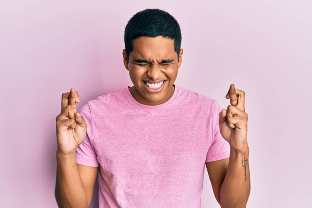 Homem hispânico bonito jovem vestindo casual rosa t-shirt gesto dedo cruzado sorrindo com esperança e olhos fechados. sorte e conceito supersticioso.  - Foto, Imagem