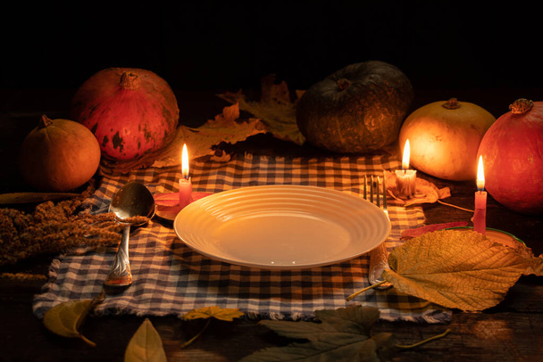 an empty plate stands on the table by candlelight on a background of pumpkin and autumn leaves, candlelit dinner, autumn table decor, festive table by candlelight - Photo, Image