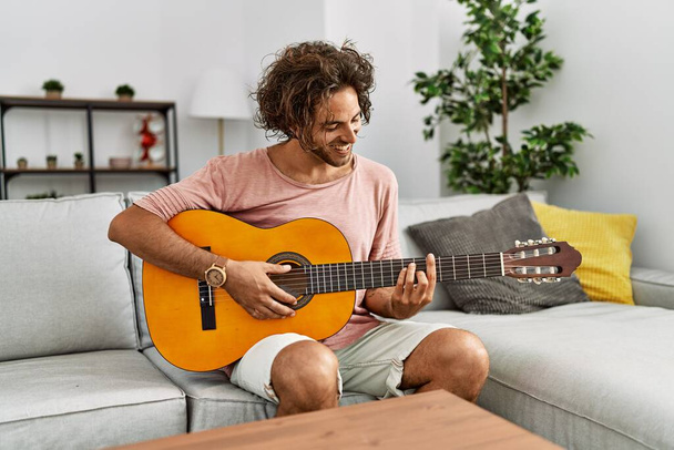 Junger hispanischer Mann lächelt glücklich und spielt klassische Gitarre zu Hause auf dem Sofa. - Foto, Bild