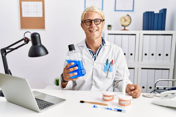 Young blond dentist man holding mouthwash at clinic looking positive and happy standing and smiling with a confident smile showing teeth  - Fotó, kép