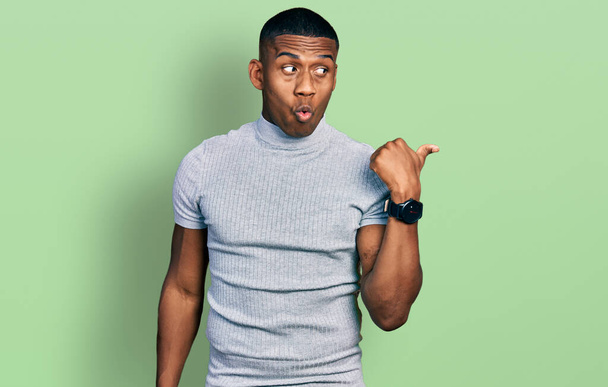Jonge zwarte man draagt casual t-shirt verrast wijzen met de hand vinger naar de zijkant, open mond verbaasd uitdrukking.  - Foto, afbeelding