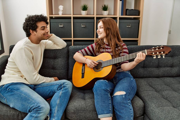 Молода пара посміхається щаслива гра на класичній гітарі, сидячи на дивані вдома
. - Фото, зображення