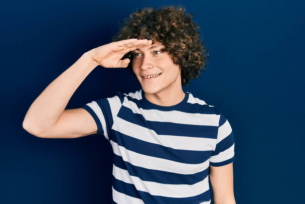 Handsome young man wearing casual striped t shirt very happy and smiling looking far away with hand over head. searching concept.  - Zdjęcie, obraz