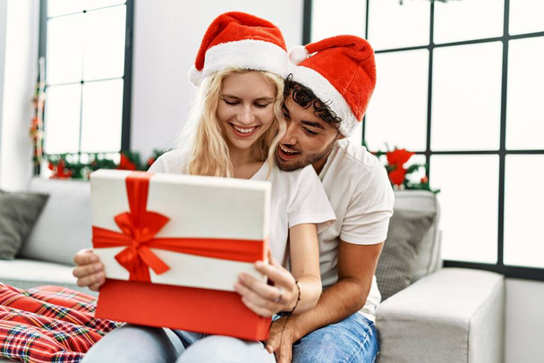 Jong stel draagt kerstmuts knuffelend en openingsgeschenk zittend op de bank thuis. - Foto, afbeelding