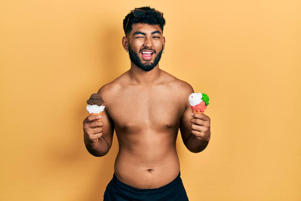 Homem árabe com barba vestindo roupa de banho comendo dois cones de sorvete piscando olhando para a câmera com expressão sexy, rosto alegre e feliz.  - Foto, Imagem
