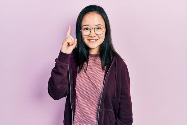 Giovane ragazza cinese indossa abiti casual e occhiali puntando il dito verso l'alto con l'idea di successo. Uscito e felice. numero uno.  - Foto, immagini