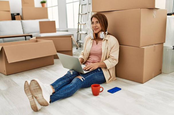 Donna caucasica di mezza età che utilizza laptop e cuffie seduti sul pavimento in una nuova casa. - Foto, immagini