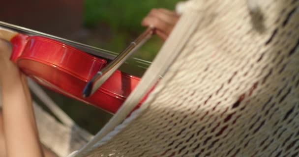 person rests in hammock in shade of trees learning to play the violin - Footage, Video