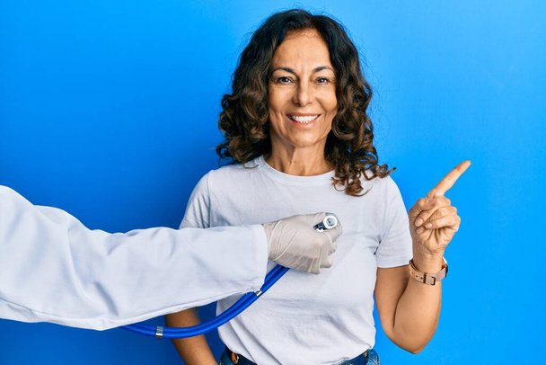 Middle age hispanic woman being auscultated by doctor smiling happy pointing with hand and finger to the side  - Fotoğraf, Görsel