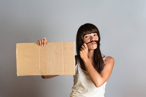 The concept of feminism and women's rights. A woman holds a cardboard banner in her hands, imitating a man's mustache on her face with her hair. Copy space. - Фото, изображение