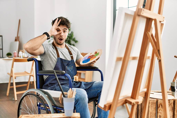 Young hispanic man sitting on wheelchair painting at art studio doing ok gesture shocked with surprised face, eye looking through fingers. unbelieving expression.  - Fotó, kép