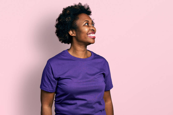 Αφροαμερικανή γυναίκα με αφρο μαλλιά φορώντας casual μωβ μπλουζάκι κοιτάζοντας μακριά στην πλευρά με χαμόγελο στο πρόσωπο, φυσική έκφραση. γελώντας σίγουρος.  - Φωτογραφία, εικόνα