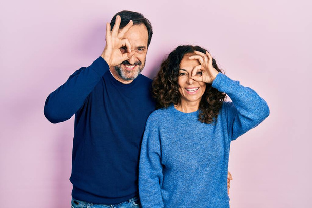 Középkorú pár spanyol nő és férfi ölelkezve és együtt állva csinál oké gesztus kézmosollyal, szem néz keresztül ujjain boldog arccal.  - Fotó, kép