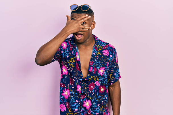 Young black man wearing hawaiian shirt and sunglasses peeking in shock covering face and eyes with hand, looking through fingers with embarrassed expression.  - Фото, зображення