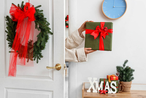 Weibliche Hände mit Weihnachtsgeschenk, verzierte weiße Tür und Kommode mit Wort XMAS nahe Lichtwand - Foto, Bild