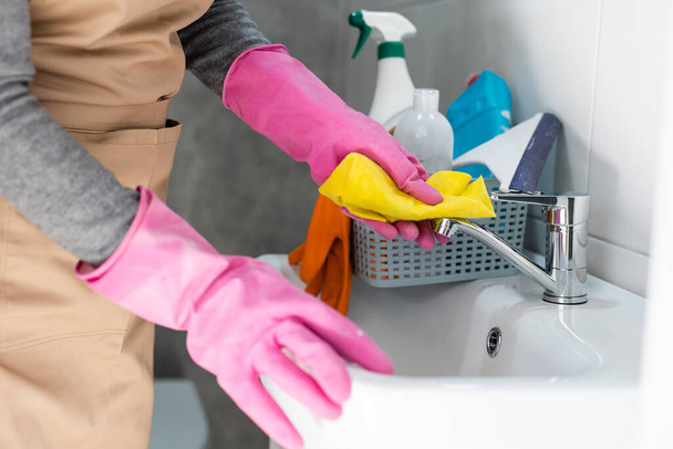 Женщина выполняет работу по дому в ванной комнате, моет раковину и кран с моющим средством
. - Фото, изображение