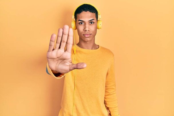 Młody, afrykański Amerykanin słuchający muzyki za pomocą słuchawek przestaje śpiewać dłonią. wyraz ostrzegawczy z negatywnym i poważnym gestem na twarzy.  - Zdjęcie, obraz