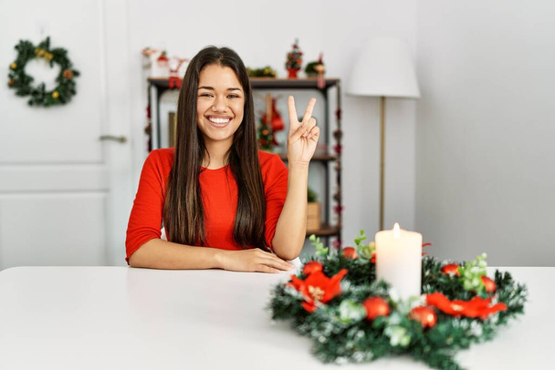 Giovane donna bruna seduta sul tavolo a Natale sorridente con la faccia felice che strizza l'occhio alla macchina fotografica facendo segno di vittoria. numero due.  - Foto, immagini