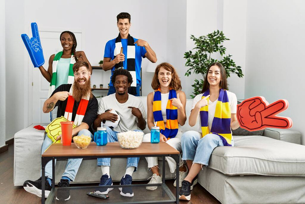 サッカーチームをサポートする友人のグループテレビを見て自宅ソファの上に座って指を指す1人の自己笑顔幸せと誇り  - 写真・画像