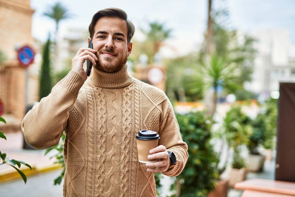 Молодой кавказский мужчина разговаривает по смартфону, пьет кофе в городе. - Фото, изображение