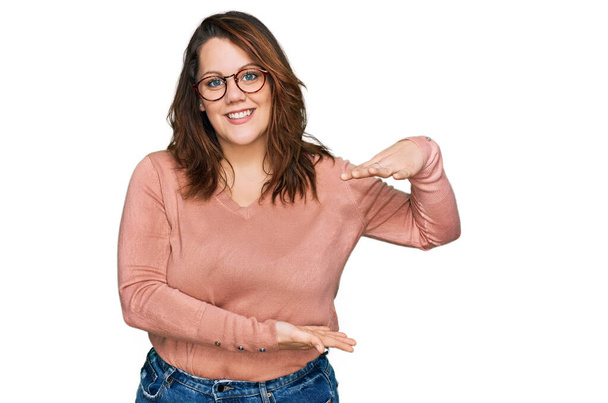 Young plus size woman wearing casual clothes and glasses gesturing with hands showing big and large size sign, measure symbol. smiling looking at the camera. measuring concept.  - Foto, Imagem