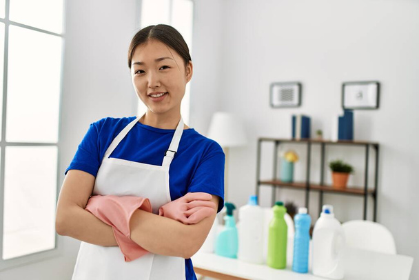 Молодая китайская домохозяйка, стоящая со скрещенными руками и моющими средствами на столе дома. - Фото, изображение