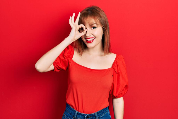 Redhead young woman wearing casual red t shirt doing ok gesture with hand smiling, eye looking through fingers with happy face.  - Foto, Bild