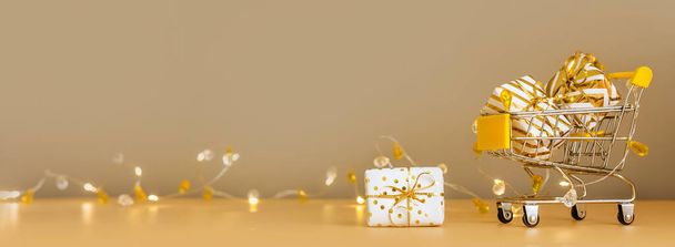 Online kerstinkopen en -verkoop. Winkelwagen met geschenkdozen op gouden achtergrond met verlichting. Supermarkt trolley vol cadeautjes. Banner met kopieerruimte - Foto, afbeelding