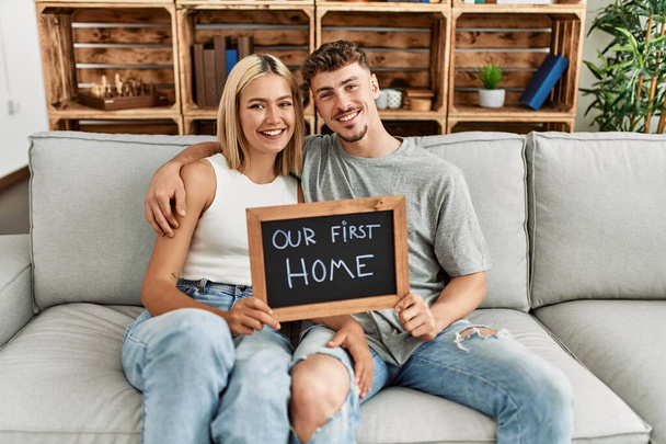 Young caucasian couple smiling happy holding blackboard with our first home message sitting on the sofa. - 写真・画像