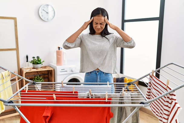 Young hispanic woman putting fresh laundry on clothesline with hand on head for pain in head because stress. suffering migraine.  - Photo, Image