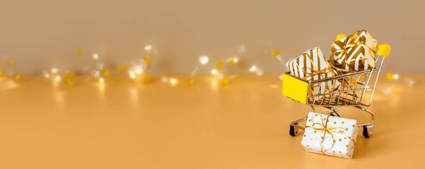 Achat et vente de Noël en ligne. Panier avec des boîtes-cadeaux sur fond doré avec des lumières. Supermarché chariot cadeaux complets. Bannière avec espace de copie - Photo, image