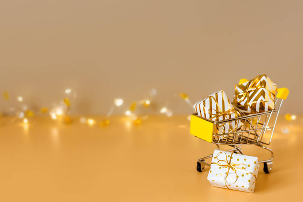 Compras y venta de Navidad en línea. Carrito de compras con cajas de regalo sobre fondo dorado con luces. Un carrito de supermercado lleno de regalos. Copiar espacio - Foto, imagen