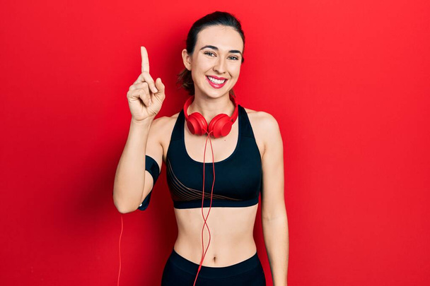 Νεαρή ισπανόφωνη κοπέλα που φοράει ρούχα γυμναστικής και χρησιμοποιεί ακουστικά που δείχνουν και δείχνουν προς τα πάνω με το δάχτυλο νούμερο ένα ενώ χαμογελά αυτοπεποίθηση και χαρούμενος.  - Φωτογραφία, εικόνα