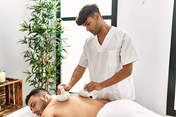 Twee Latijns-Amerikaanse mannen fysiotherapeut en patiënt masseren terug met behulp van thai zakken bij beauty center - Foto, afbeelding