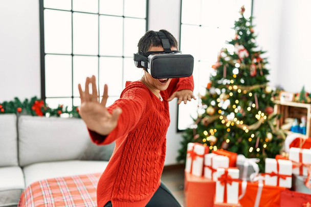 Young hispanic woman playing video game using vr glasses standing by christmas tree at home - 写真・画像