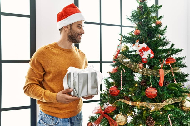 Junger Araber lächelt glücklich und hält Geschenk am heimischen Weihnachtsbaum. - Foto, Bild