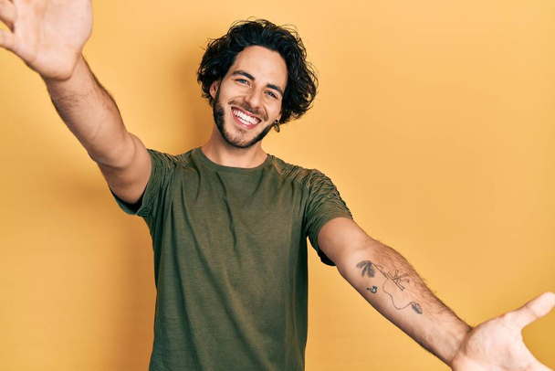 Schöner hispanischer Mann in lässigem grünem T-Shirt, der lächelnd mit offenen Armen in die Kamera blickt, um sich zu umarmen. fröhlicher Ausdruck, der das Glück umarmt.  - Foto, Bild