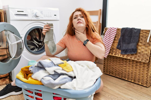Молодая рыжая женщина кладет грязное белье в стиральную машину касаясь болезненной шеи, боль в горле от гриппа, заноз и инфекции  - Фото, изображение