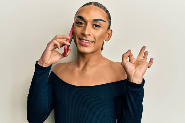 Hispanic transgender man wearing make up and long hair having conversation talking on the smartphone doing ok sign with fingers, smiling friendly gesturing excellent symbol  - Valokuva, kuva
