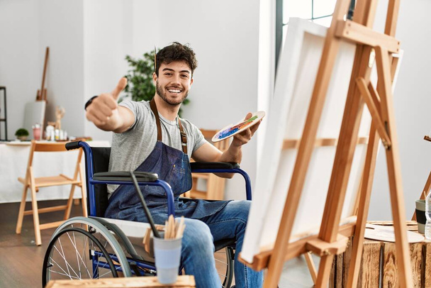 Νεαρός Ισπανόφωνος κάθεται σε αναπηρική καρέκλα ζωγραφική στο στούντιο τέχνης έγκριση κάνει θετική χειρονομία με το χέρι, τους αντίχειρες επάνω χαμογελώντας και χαρούμενος για την επιτυχία. νικήτρια χειρονομία.  - Φωτογραφία, εικόνα