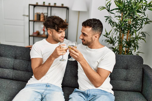 Dos hombres hispanos brindando en pareja con una copa de vino sentados en un sofá en casa - Foto, imagen