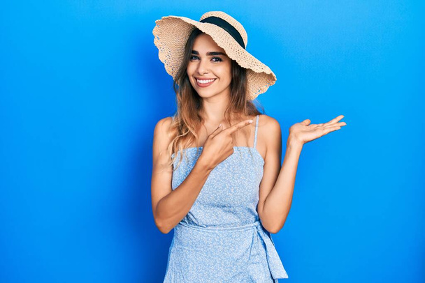 Νεαρή ισπανόφωνη κοπέλα φορώντας καλοκαιρινό καπέλο κατάπληκτη και χαμογελώντας στην κάμερα ενώ παρουσιάζει με το χέρι και δείχνοντας με το δάχτυλο.  - Φωτογραφία, εικόνα