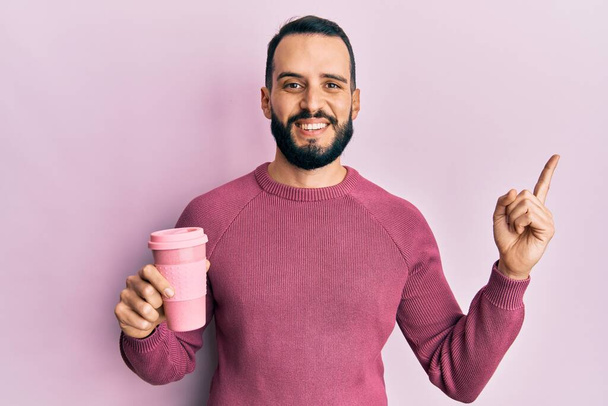 Young man with beard drinking a take away cup of coffee smiling happy pointing with hand and finger to the side  - Foto, immagini