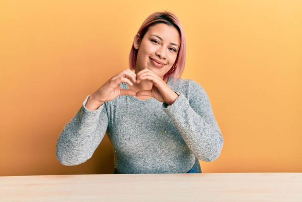 Hispanic woman with pink hair wearing casual clothes sitting on the table smiling in love showing heart symbol and shape with hands. romantic concept.  - Photo, Image