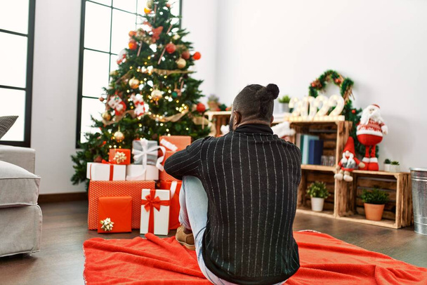 Young african american man on back view sitting on the floor looking christmas tree at home - Foto, Bild
